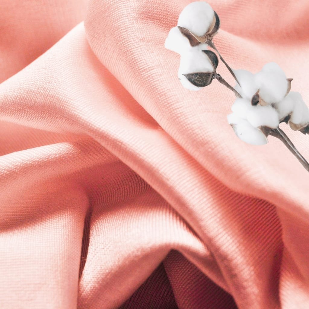 A close-up image of soft, peach-colored mercerized cotton fabric with a delicate cotton branch resting on top, highlighting the fabric’s smooth, shiny, and slightly textured surface.