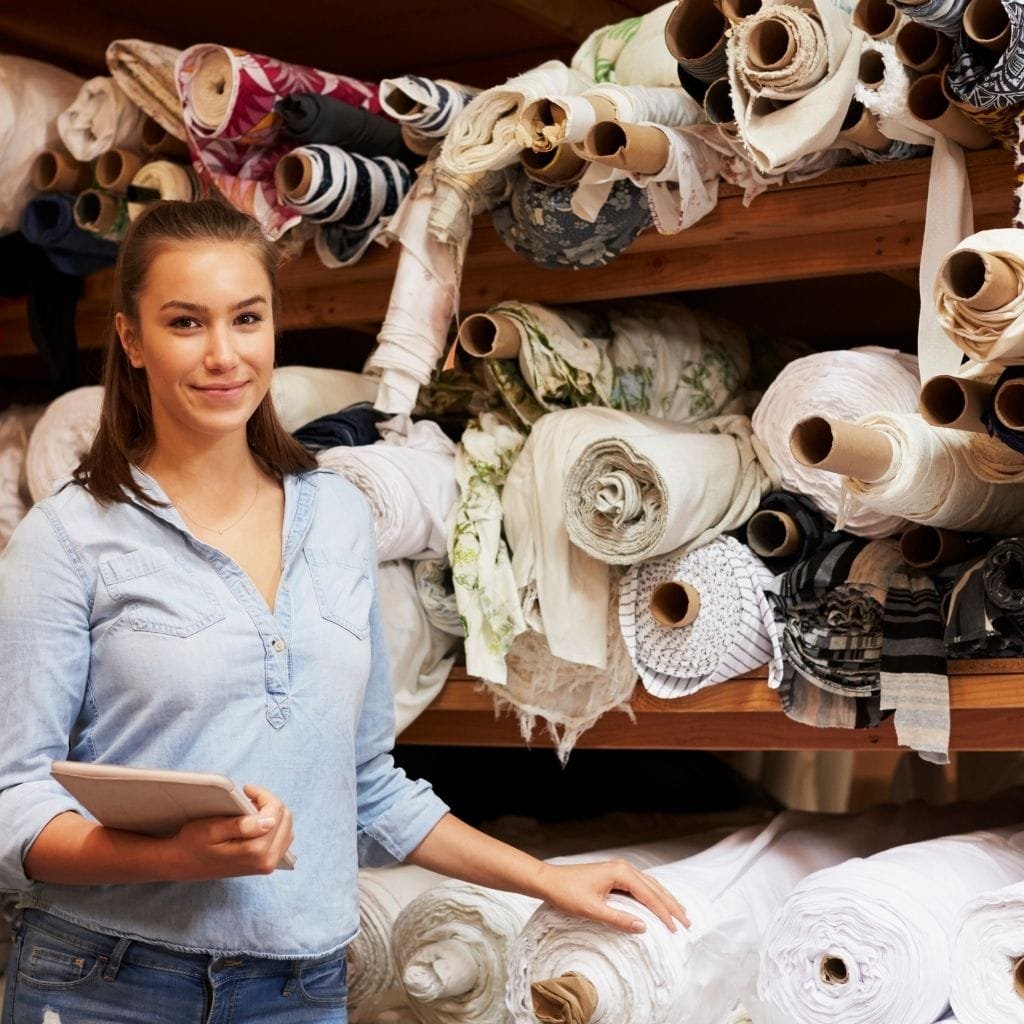  A young woman smiling at the camera while holding a notebook, standing in front of shelves stacked with rolls of fabric and textiles in various colors and patterns.