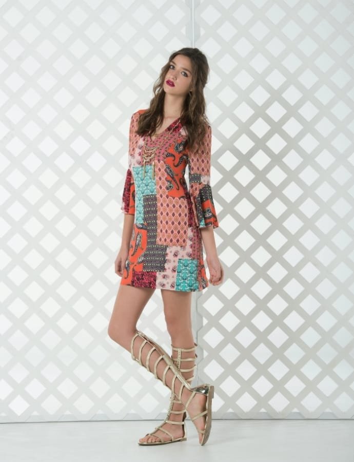 A young woman with long wavy hair and red lipstick, wearing a colorful patchwork dress with mixed patterns, paired with gladiator sandals, standing against a white geometric lattice background.