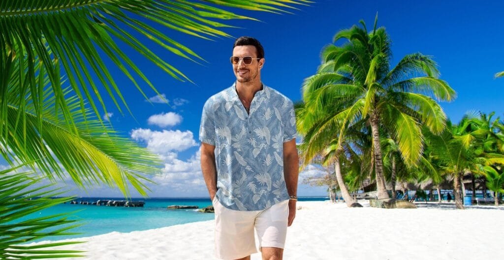 A man walking along a tropical beach with white sand, turquoise water, and lush green palm trees, wearing a light blue Hawaiian shirt with white floral patterns and white shorts