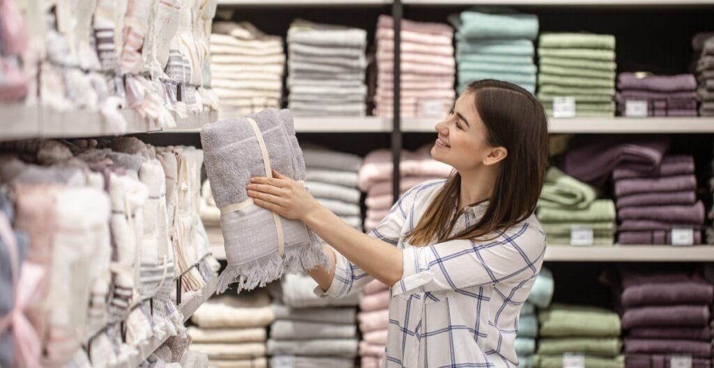 A woman shopping for towels in a store, holding and examining a folded grey towel with a smile on her face.