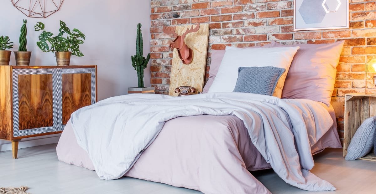 A cozy bedroom with a bed covered in light pink and white bedding, set against a brick wall with decorative elements, including a cactus plant, a wooden side table, and framed art.