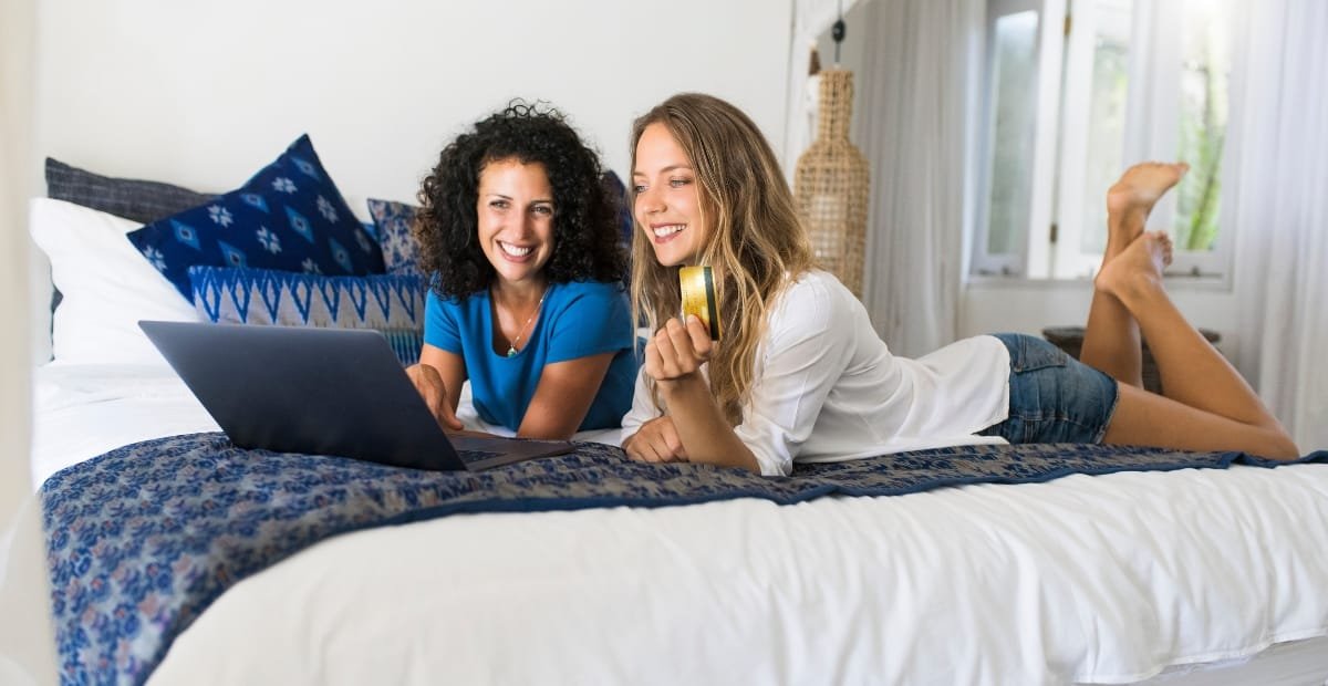 Two women lying on a bed, smiling and looking at a laptop, with one holding a roll of tape.