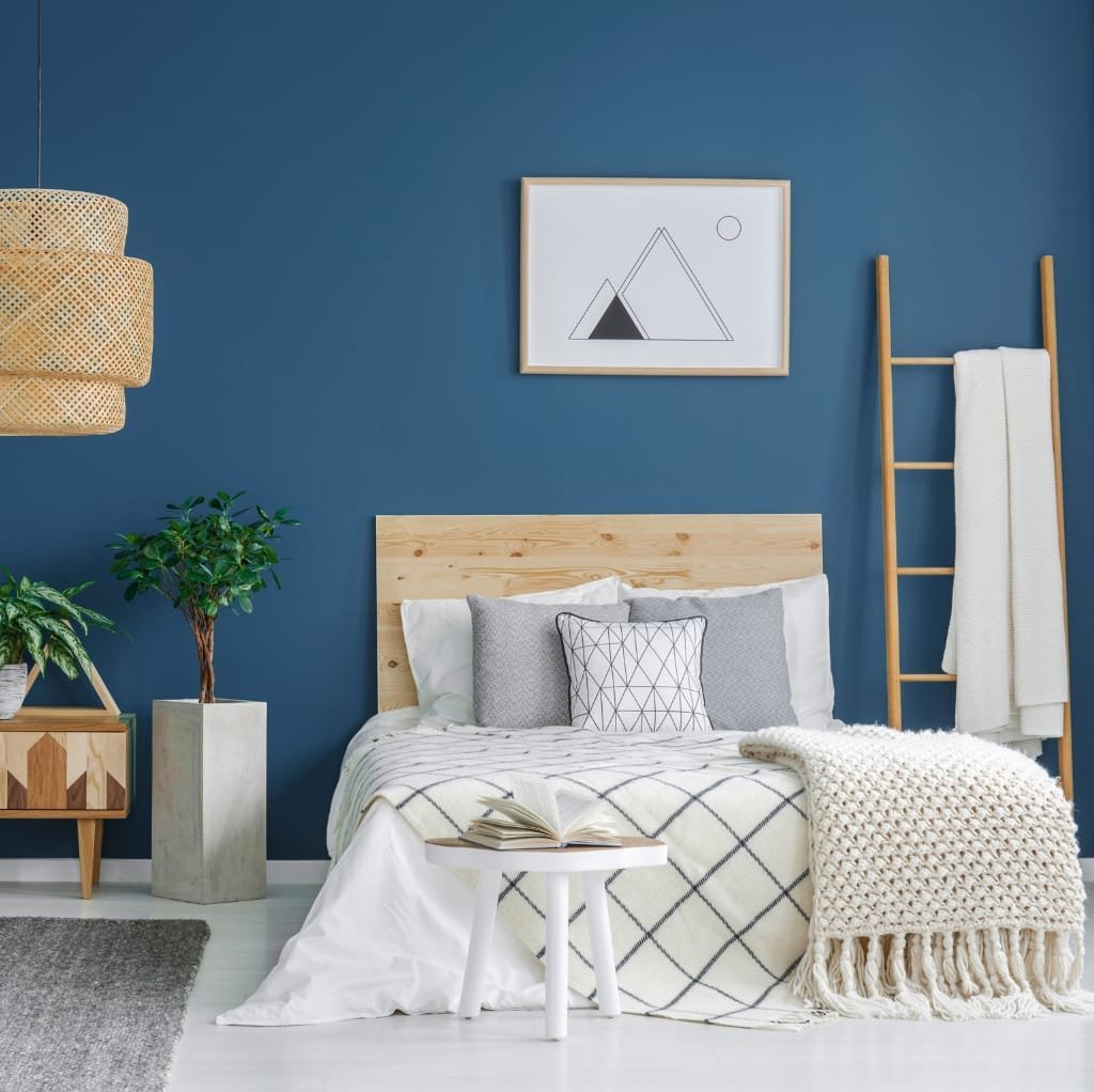 A cozy bedroom featuring a minimalist design with a blue accent wall, a wooden headboard, a small white side table with an open book, a textured cream throw blanket on the bed, decorative pillows, a tall wooden ladder with a white towel, a round woven pendant light, a potted plant on a wooden nightstand, and simple geometric wall art.