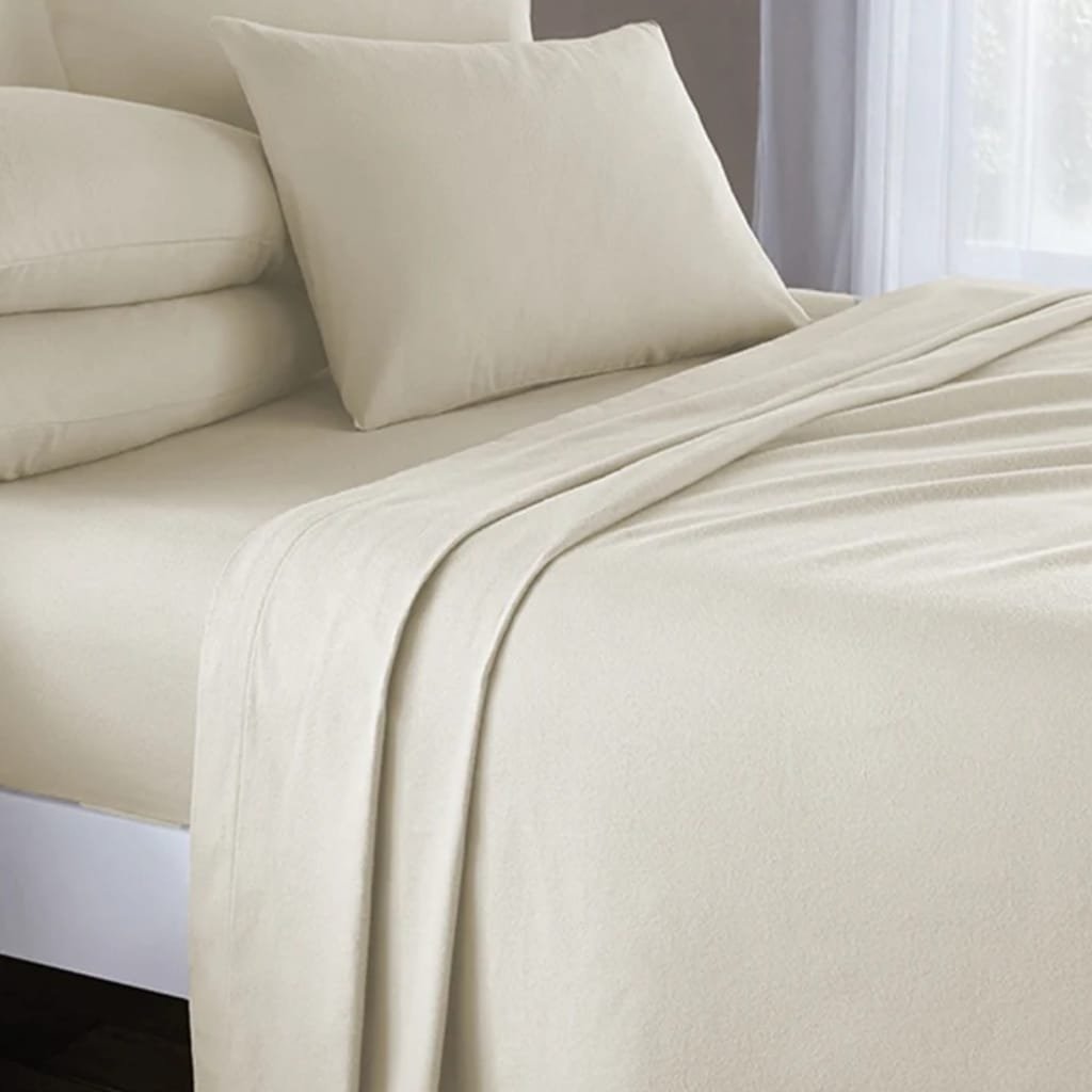A neatly made bed with cream-colored sheets, two matching pillows, and a white bed frame. The bed is positioned near a window with sheer curtains, allowing natural light to softly illuminate the room.