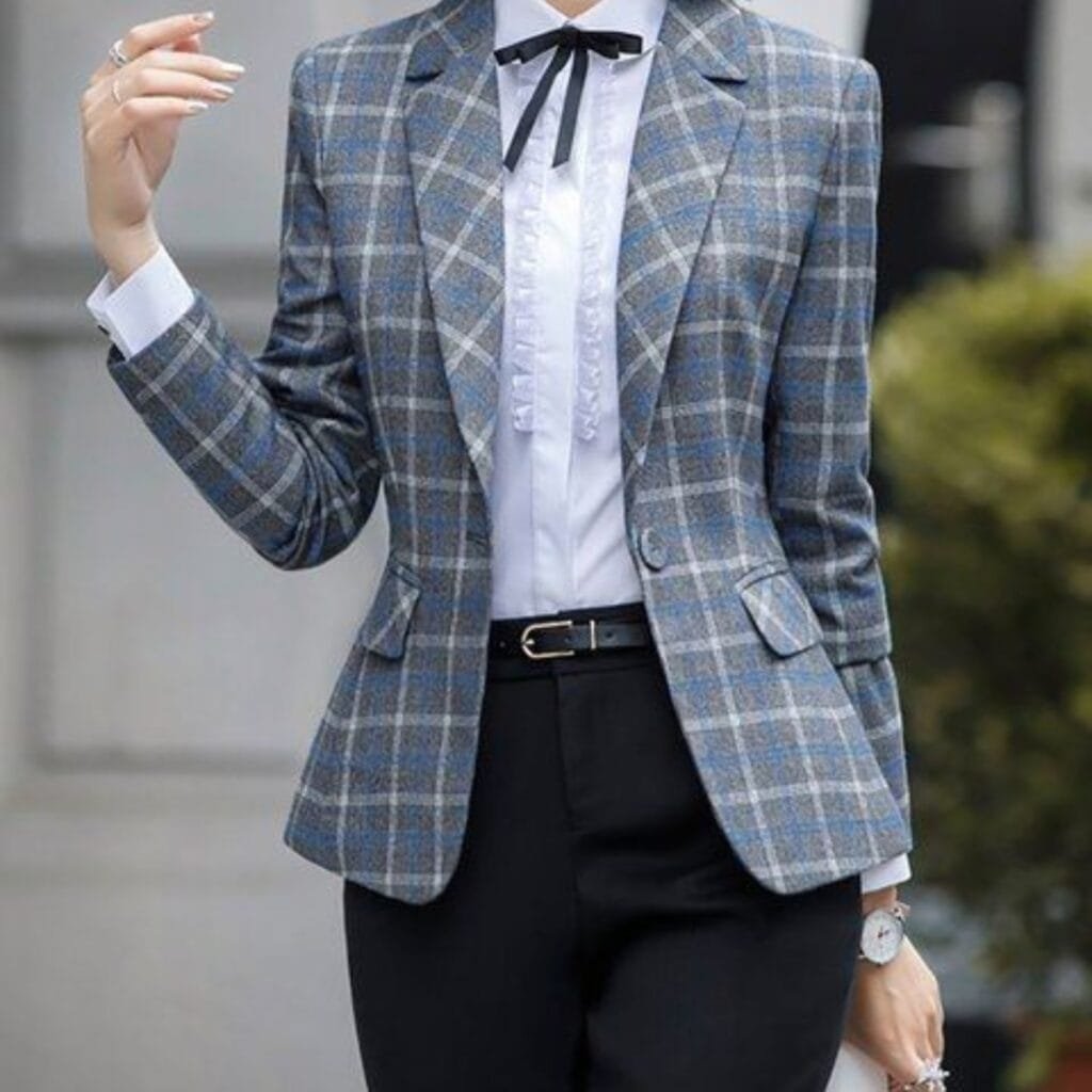 A woman wearing a stylish plaid blazer over a white blouse with a black bow tie, paired with black trousers, standing outdoors.