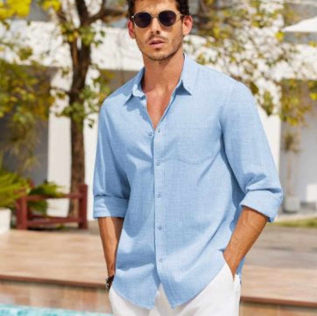 A stylish man wearing a light blue, long-sleeved, button-down shirt with rolled-up sleeves, paired with white pants, standing outdoors with greenery in the background. COOFANDY Men’s Shirts