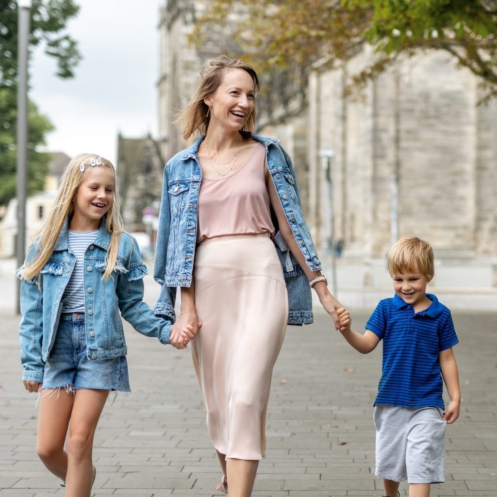 A smiling woman walking outdoors holding hands with a young girl and a young boy, all dressed in casual denim outfits, with trees and historic buildings in the background.