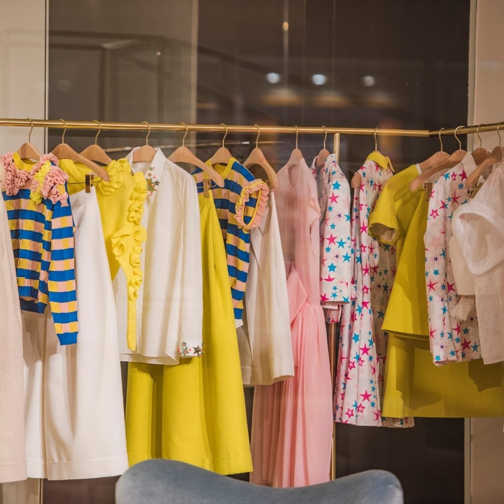 A display of women's clothing hanging on a gold-colored rack, featuring pastel and colorful dresses and blouses with various patterns including stars, stripes, and ruffles.