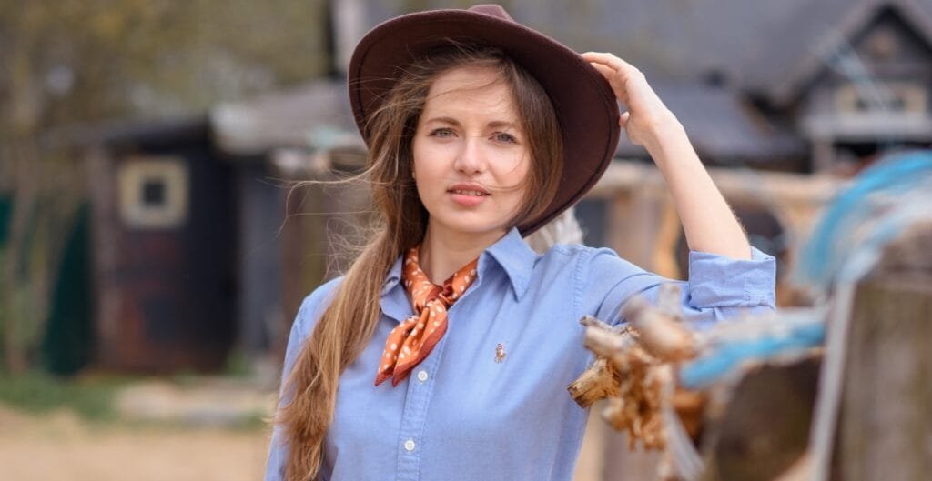 A woman wearing a light blue chambray shirt, a brown wide-brim hat, and an orange polka-dot scarf around her neck, standing outdoors.
