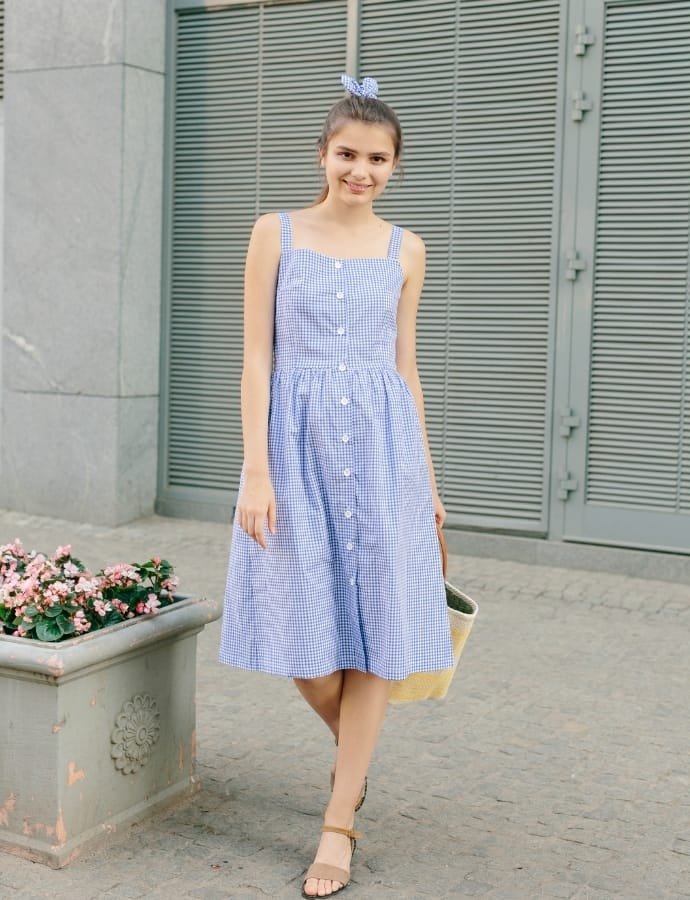 Young woman wearing a blue and white checkered cotton babydoll dress with spaghetti straps, standing outdoors near a pot of pink flowers and gray metal shutters, smiling and holding a beige tote bag.