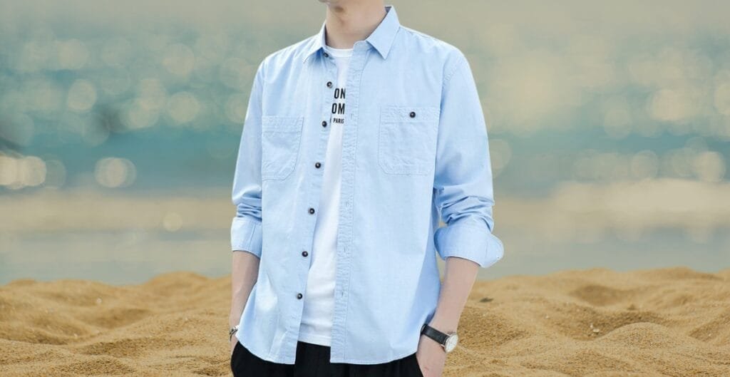 A person standing on a sandy beach, wearing a light blue cotton button-down shirt over a white t-shirt, with rolled-up sleeves.