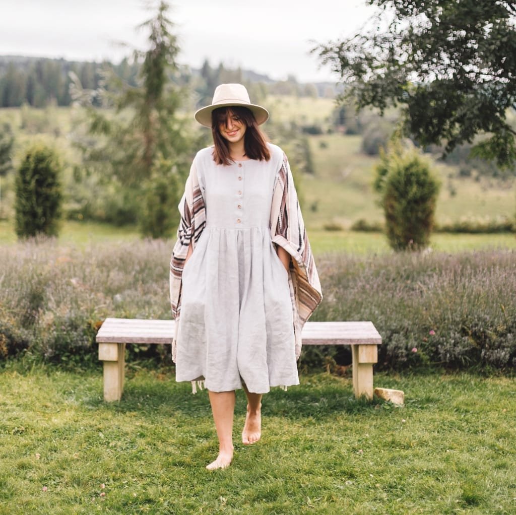 A woman walking barefoot on a grassy field, wearing a light-colored, loose-fitting cotton kaftan dress with a plaid shawl and a wide-brimmed hat, surrounded by greenery and a blurred natural landscape.