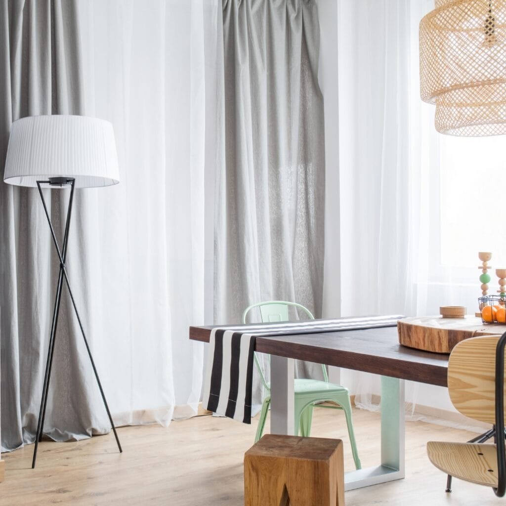 A modern interior scene featuring a wooden table with a light-colored wooden block on top, surrounded by various chairs, including a mint green metal chair and a wooden chair. White and gray curtains hang in the background, with a tall, minimalist black tripod floor lamp on the left and a woven pendant light hanging from the ceiling on the right.