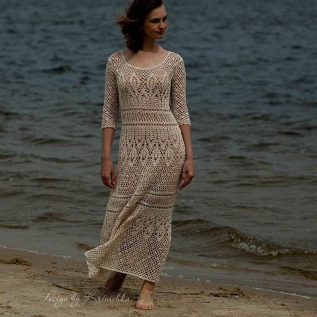  A woman in a long, beige crochet lace dress walks barefoot along the shoreline at the beach, with the ocean in the background.