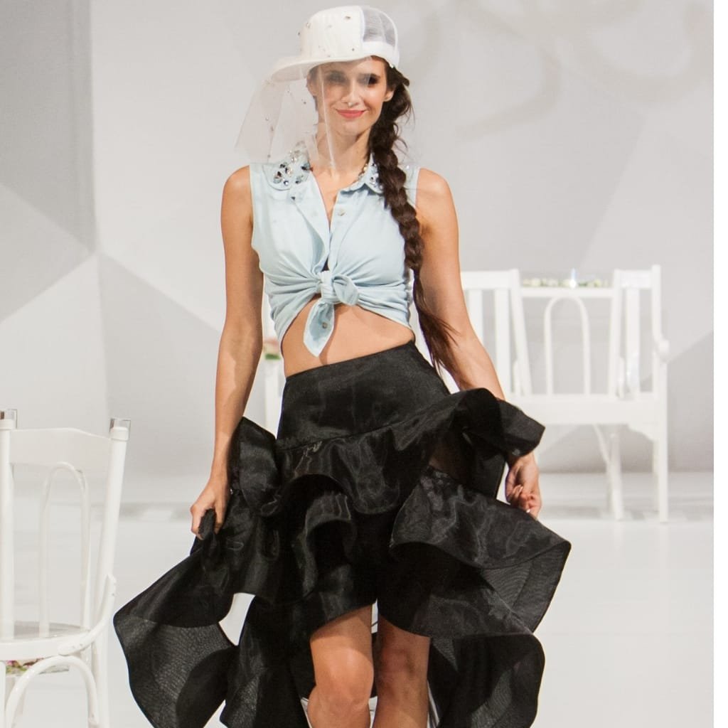 A woman walking confidently on the runway in a fashion show, wearing a sleeveless knotted top and a voluminous black skirt, with a clear face shield and cap, demonstrating the use of Crystal Organza fabric in the Catwalk Secret collection.