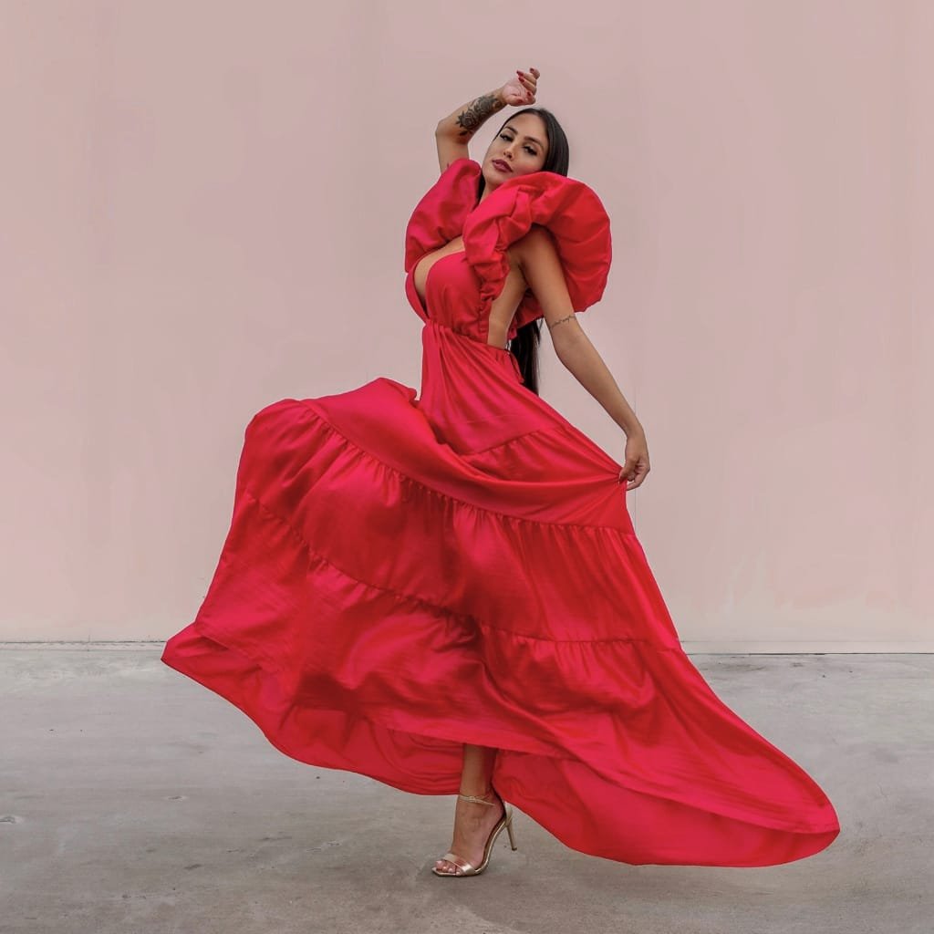 A woman wearing a flowing, vibrant red evening gown made from crystal organza fabric. She is posing gracefully against a soft pink background, showcasing the fabric’s elegant drape and shimmer.