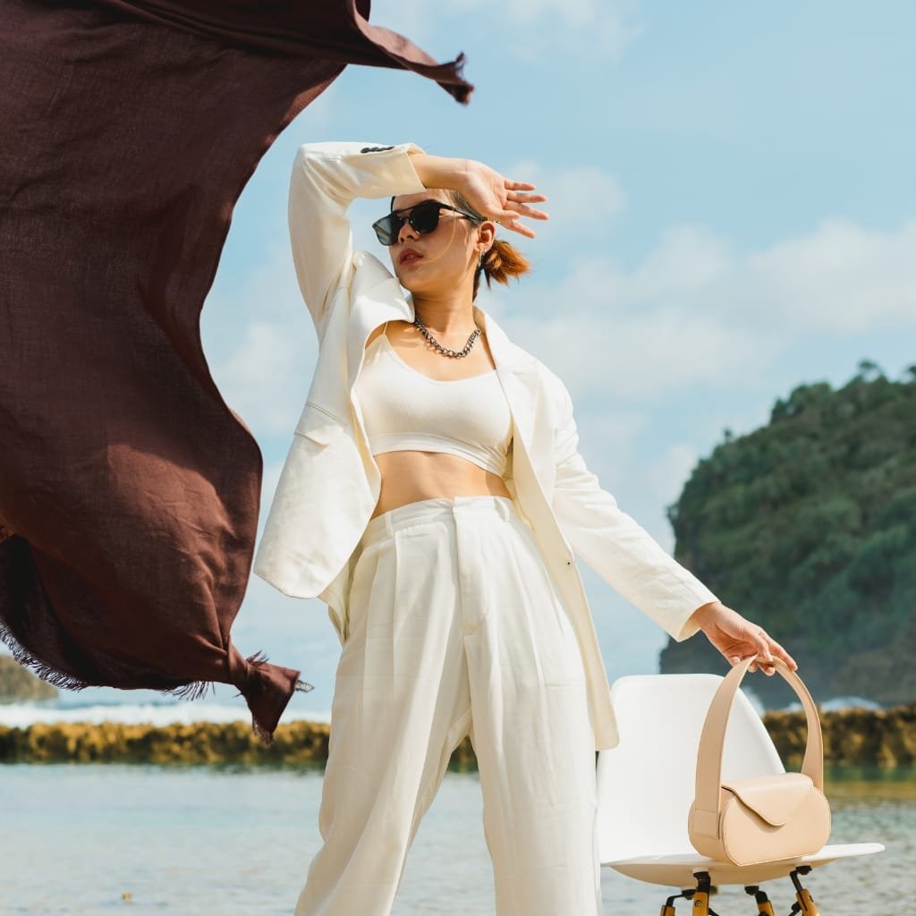 A woman in a stylish beige outfit with sunglasses, standing on a beach, holding a beige handbag, with a large brown scarf billowing in the wind.
