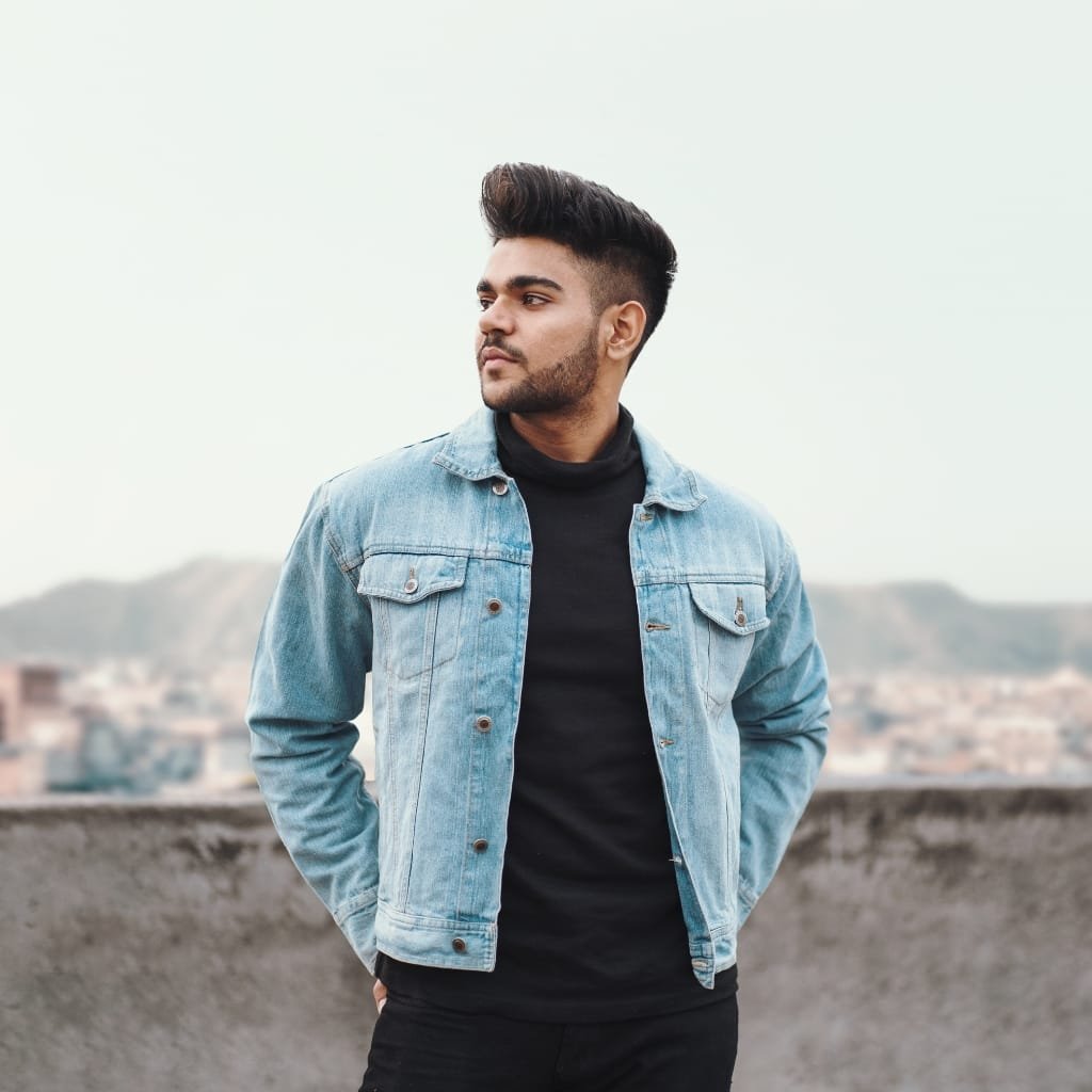A young man stands outdoors on a rooftop, wearing a light blue denim chambray jacket over a black turtleneck, with a cityscape blurred in the background.