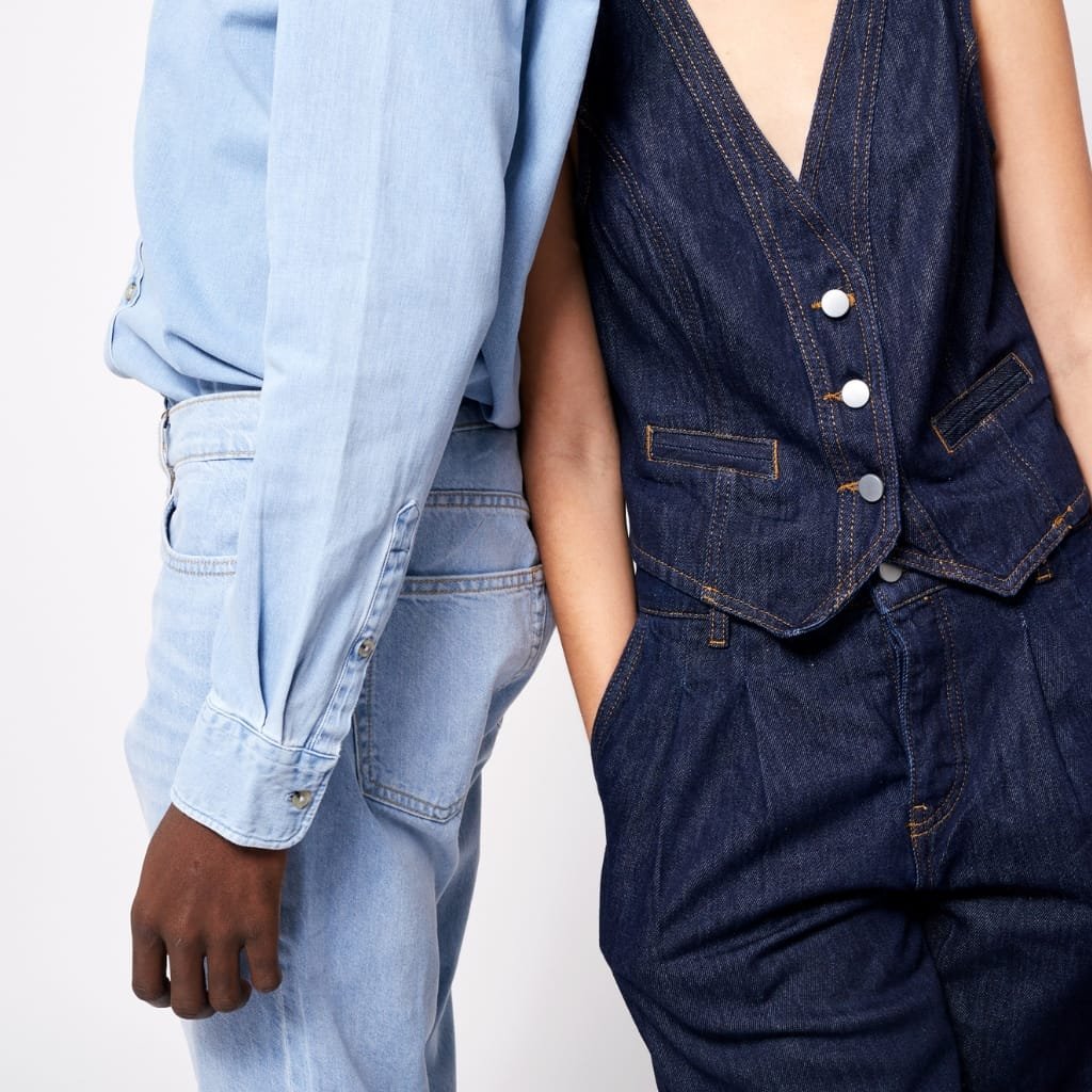 Close-up of two people wearing denim outfits: one in a light blue denim shirt and pants, and the other in a dark blue denim sleeveless vest and pants, showcasing a styling for all genders.