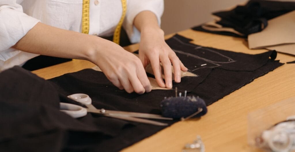 A person is working on a sewing project, cutting fabric with scissors on a table, surrounded by sewing tools.