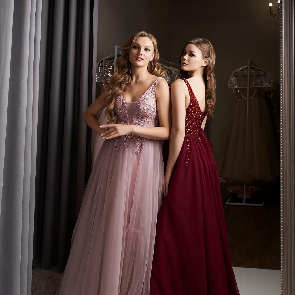 Two women in elegant evening gowns standing in front of a mirror, one in a light pink organza silk gown with intricate embroidery and the other in a deep red organza silk gown with embellished details.
