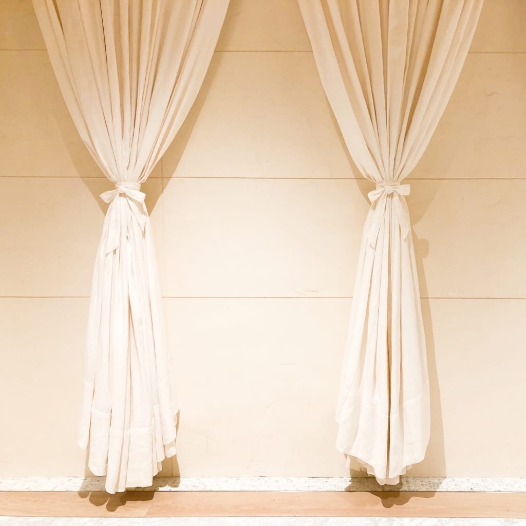 Cream-colored curtains tied back in front of a plain beige tiled wall.