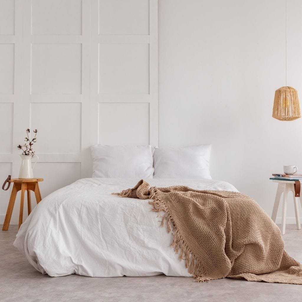 A minimalist bedroom with a white bed, two white pillows, and a beige textured throw blanket. On either side of the bed are small wooden side tables—one with a vase of dried cotton stems and the other with a cup and books. A woven pendant light hangs on the right, and the background features a plain, light-colored wall.