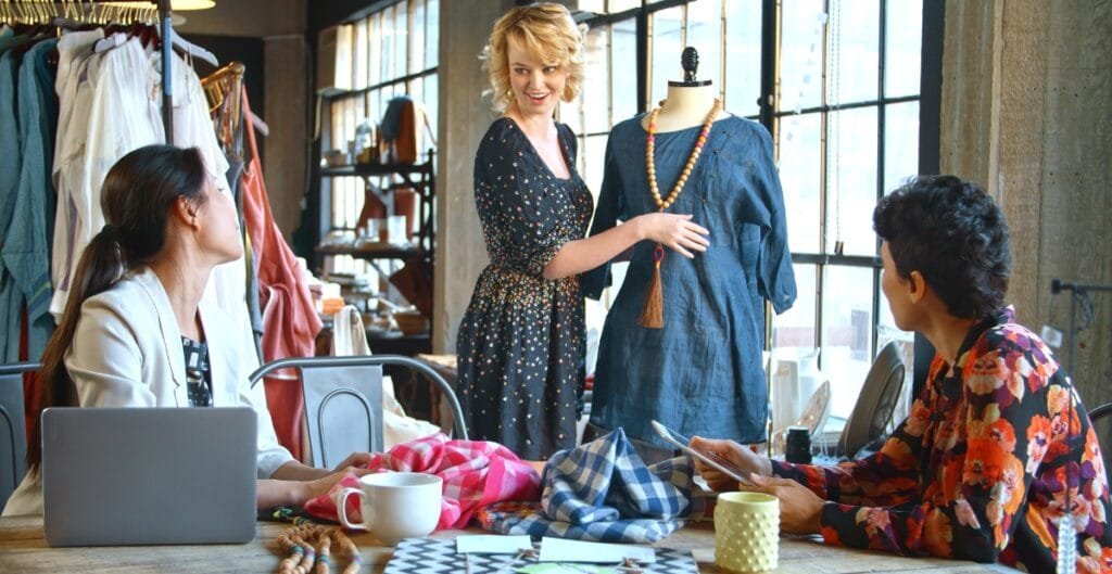 Three women are engaged in a discussion inside a boutique or clothing store. One woman is standing and showing a blue dress on a mannequin, while the other two women are seated at a table, looking at her and a magazine.
