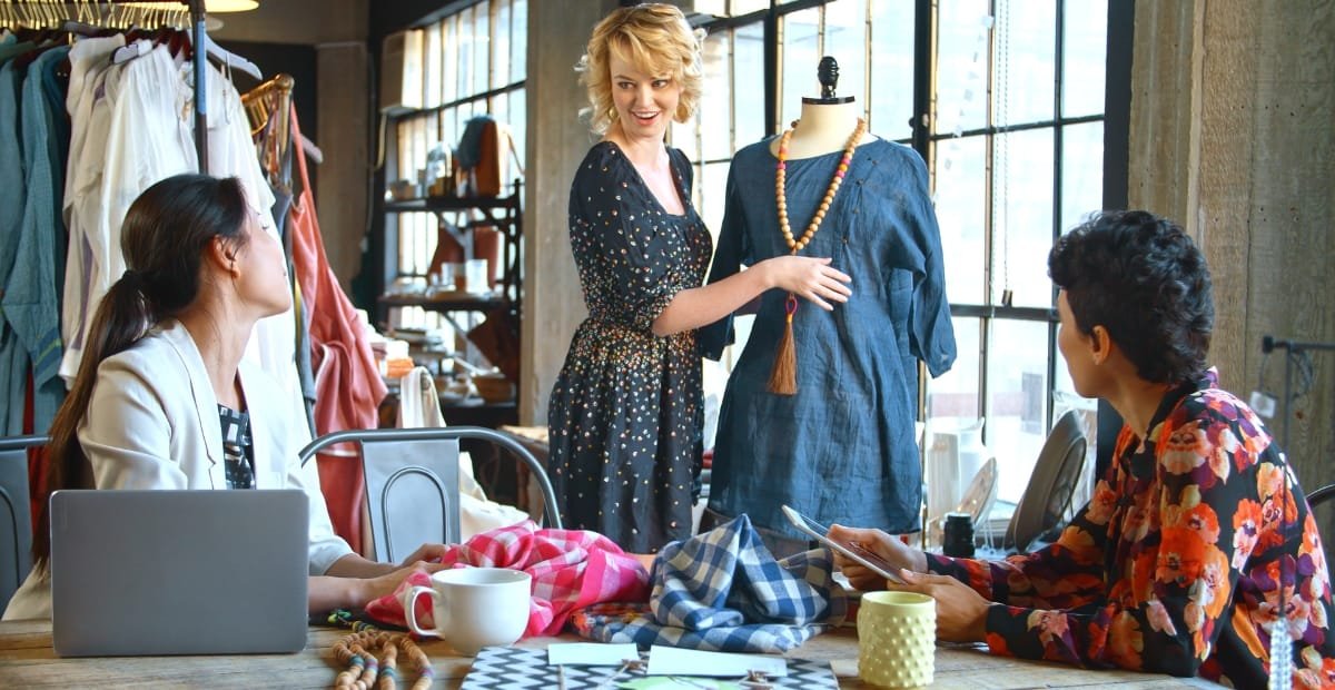 Three women are engaged in a discussion inside a boutique or clothing store. One woman is standing and showing a blue dress on a mannequin, while the other two women are seated at a table, looking at her and a magazine.