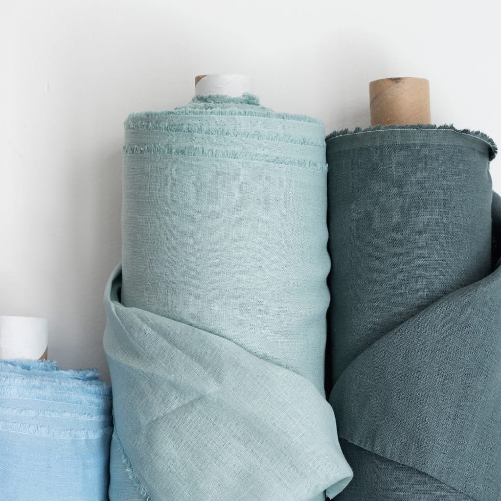  Several rolls of fabric in shades of pale blue and gray, standing upright against a plain white wall.
