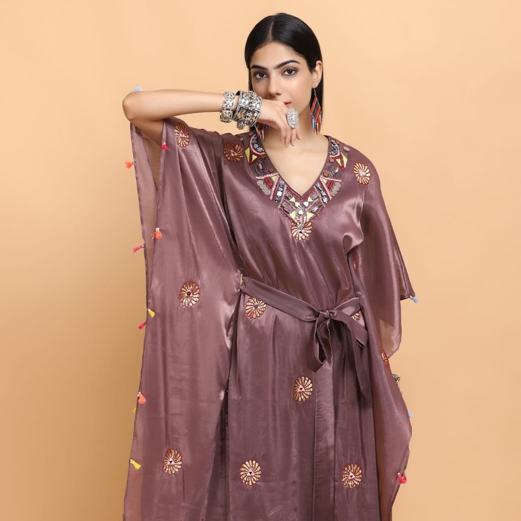 A woman wearing a shiny, embroidered satin organza dress with colorful embellishments around the neckline, accessorized with silver jewelry and multicolored earrings, stands against a plain beige background.