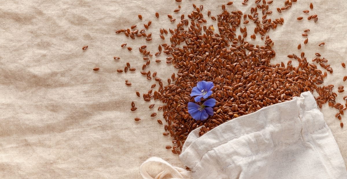Brown flax seeds spilling out of a white cloth bag on a beige surface, with two small blue flowers resting among the seeds.