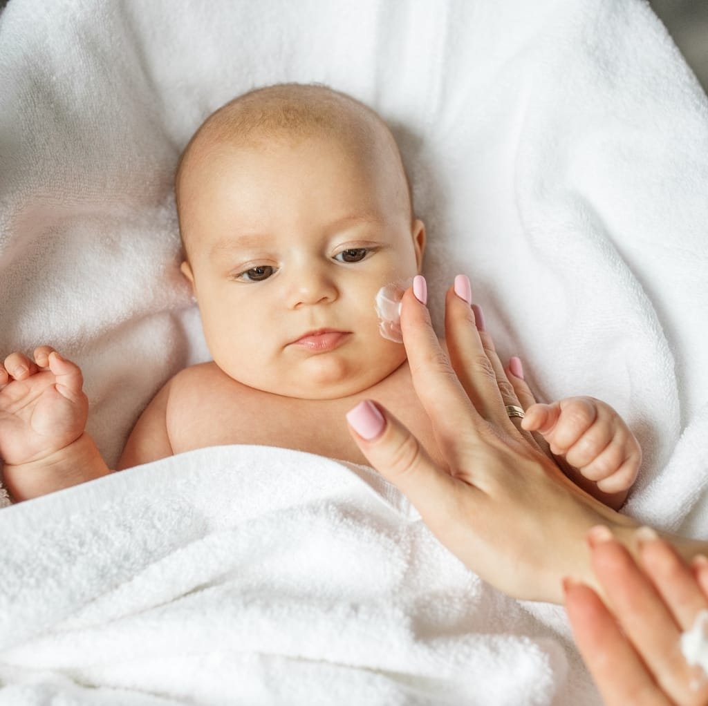 A baby with delicate skin lying on a soft white towel while a person applies gentle baby lotion or cream on their cheek.