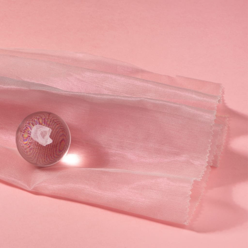 A piece of light pink crystal organza fabric draped on a soft pink background, with a glass marble reflecting intricate rainbow-colored patterns placed nearby.