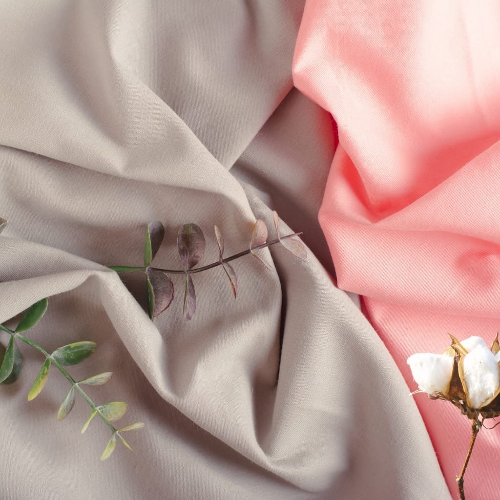 Close-up image of soft, draped fabric in beige and pink tones with small sprigs of leaves and a cotton flower