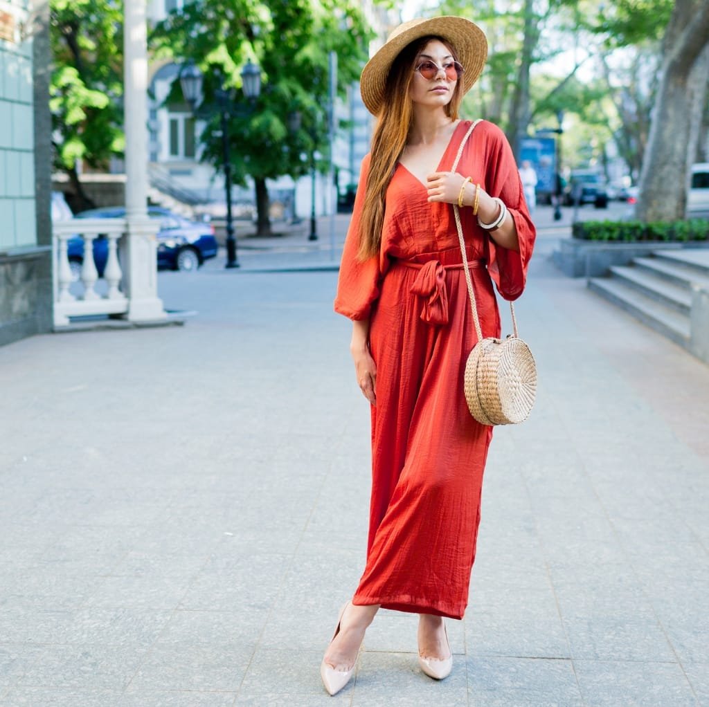 A woman wearing a stylish, flowing red maxi cotton dress with a wide-brim straw hat, sunglasses, and a woven round handbag, standing on a city sidewalk with greenery in the background.