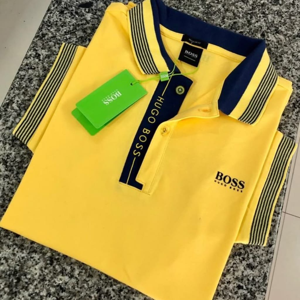 A bright yellow Hugo Boss polo shirt with navy accents, neatly folded on a granite countertop. The shirt features a navy collar with yellow and navy striped detailing, a button placket with "HUGO BOSS" embroidered in yellow on the navy strip, and the "BOSS" logo on the chest.