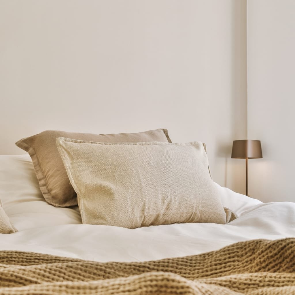 A neatly made bed with beige pillows and a textured blanket, beside a modern bedside lamp in a cozy, neutral-colored bedroom.