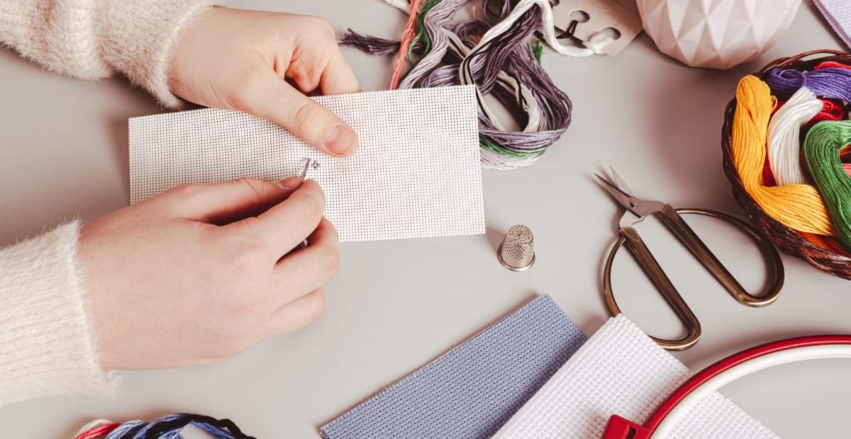 A person stitching miniature canvas on a table surrounded by cross-stitch supplies, including colorful embroidery threads, scissors, a thimble, and fabric.