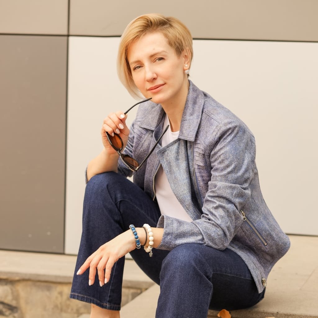 A woman with short blonde hair, wearing a gray denim jacket and dark jeans, sits on a step outdoors. She holds sunglasses in one hand and looks at the camera with a relaxed expression.