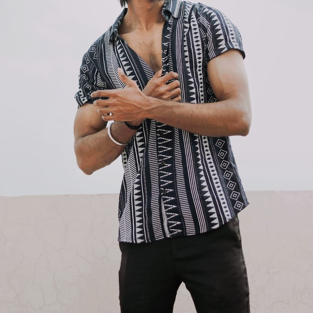A man wearing a stylish, short-sleeved summer shirt with a black and white geometric tribal pattern, paired with black pants, standing against a plain light background.