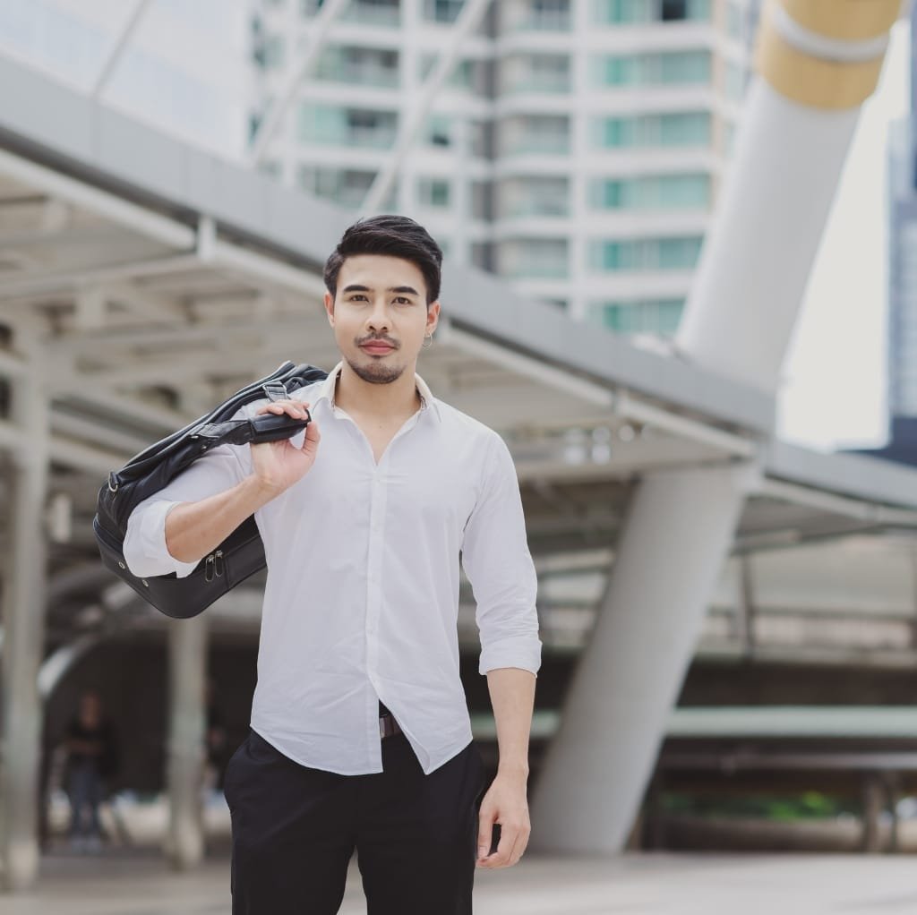 A young man dressed in a white linen shirt, holding a black bag over his shoulder, standing outdoors in an urban setting.
