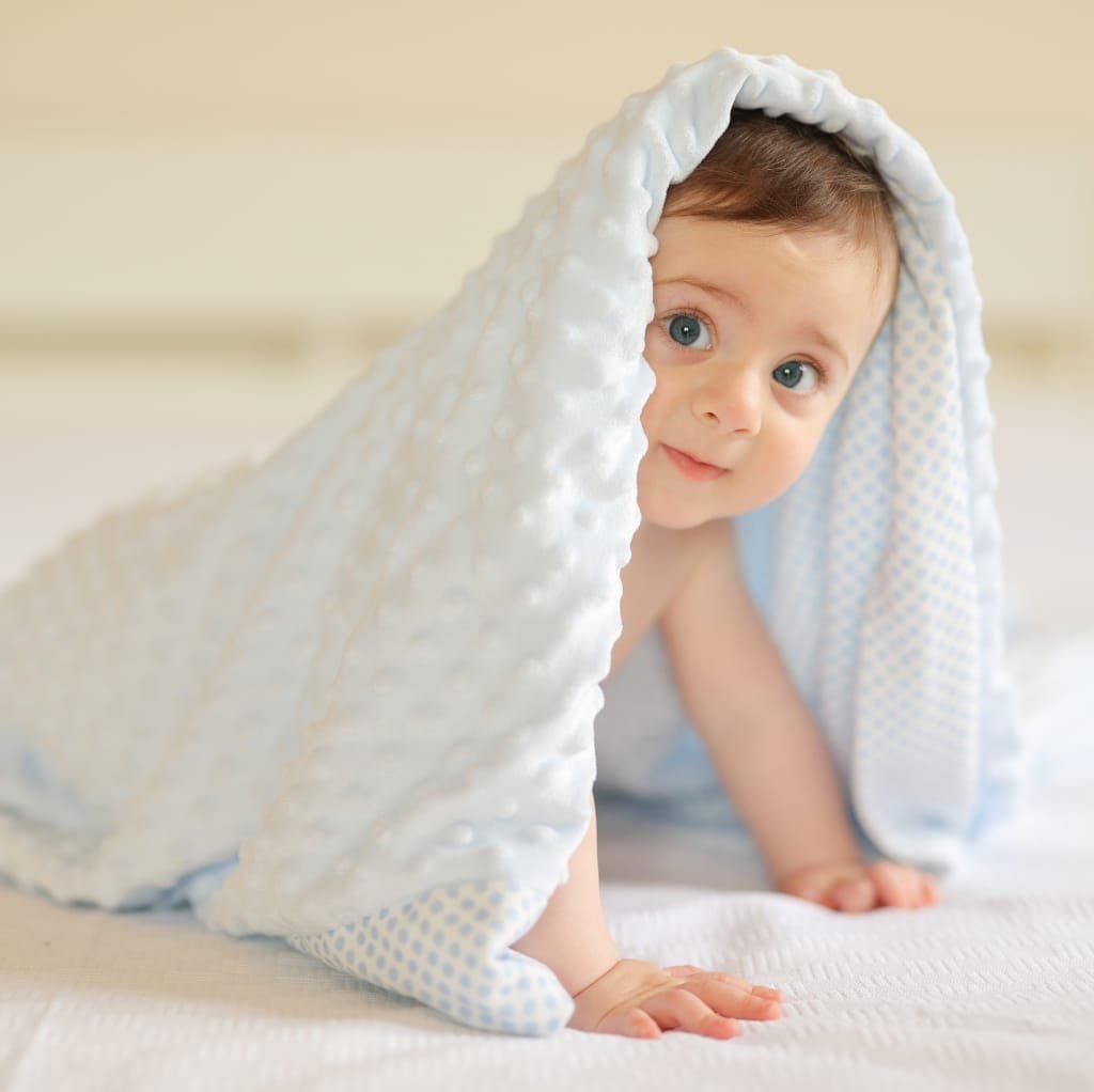 A baby crawling on a white surface, partially covered with a soft, plush blanket that drapes over their head, with bright blue eyes and a gentle expression.