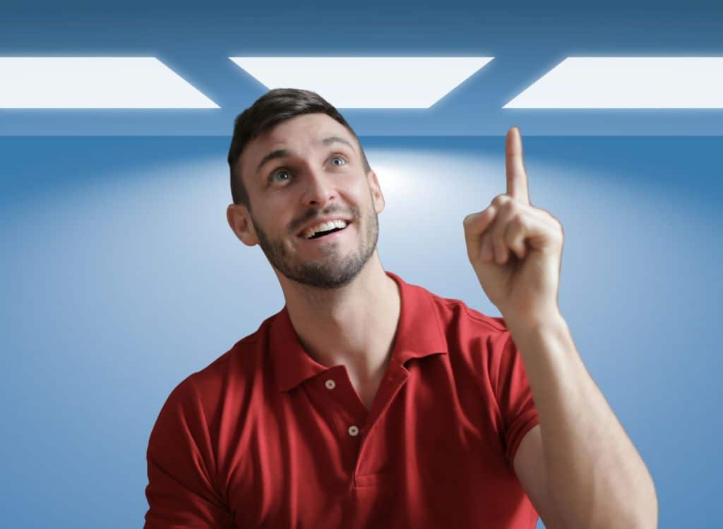 A smiling man pointing upwards, standing in front of a modern ceiling light, wearing a red polo shirt.