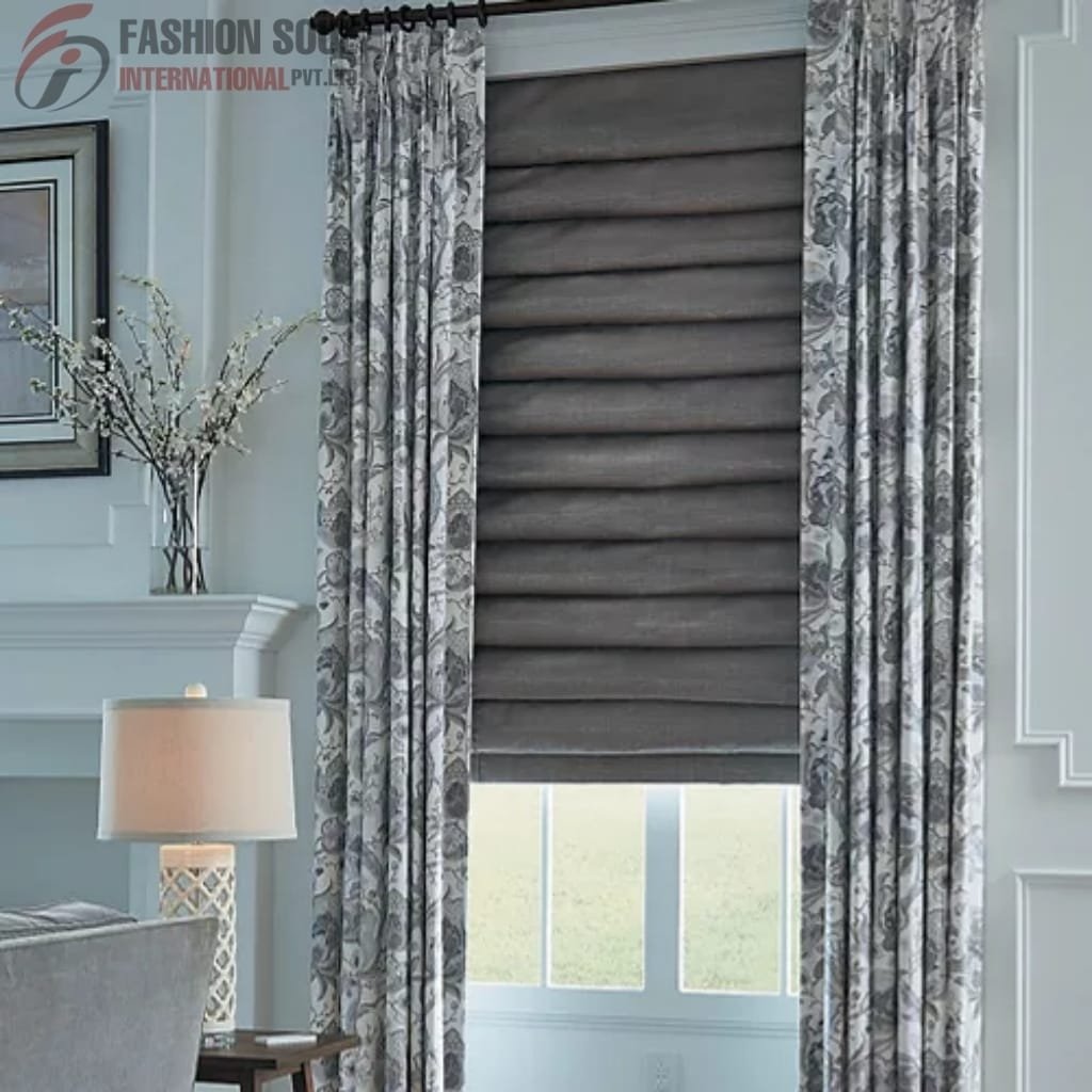 A window with a grey Roman shade partially pulled down, framed by floral-patterned curtains in shades of grey and white. To the left, there is a table lamp with a white lampshade, a side table, and a decorative branch arrangement on a mantelpiece in a cozy, well-decorated room.