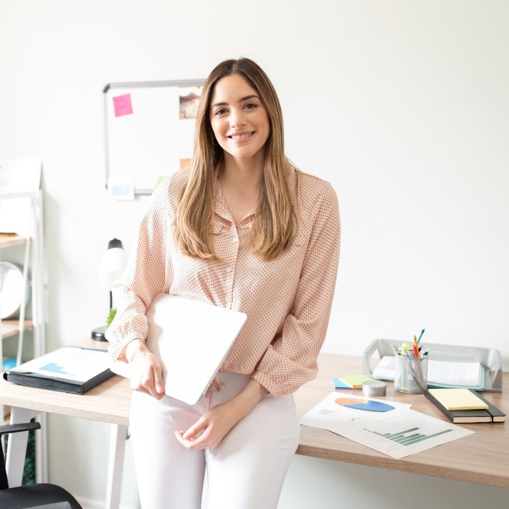 Smiling woman with long, straight brown hair in office attire holding a laptop, standing next to a desk with office supplies, charts, and documents, in a bright, modern office space.