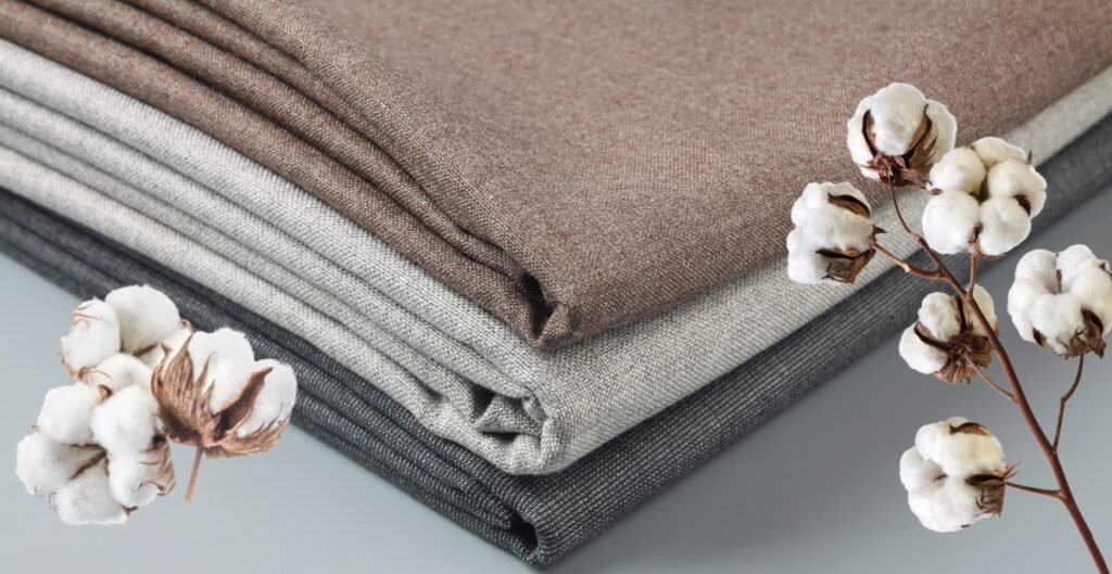 Three neatly folded pieces of fabric in shades of brown, beige, and gray with cotton branches placed beside them.