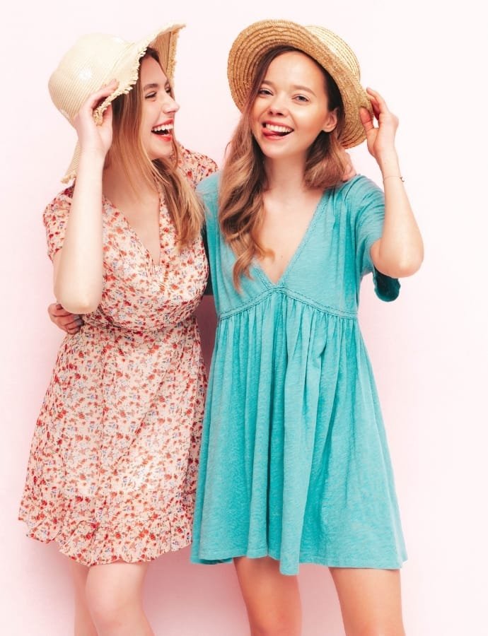 Two young women smiling and wearing summer dresses with straw hats against a light pink background.