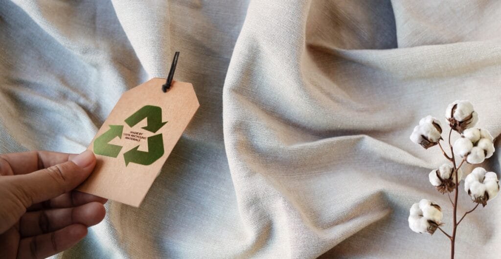A close-up of a hand holding a beige clothing tag with a green recycling symbol and the text "MADE BY RECYCLE.ME" on a piece of light-colored fabric, with a small branch of cotton flowers resting on the fabric.
